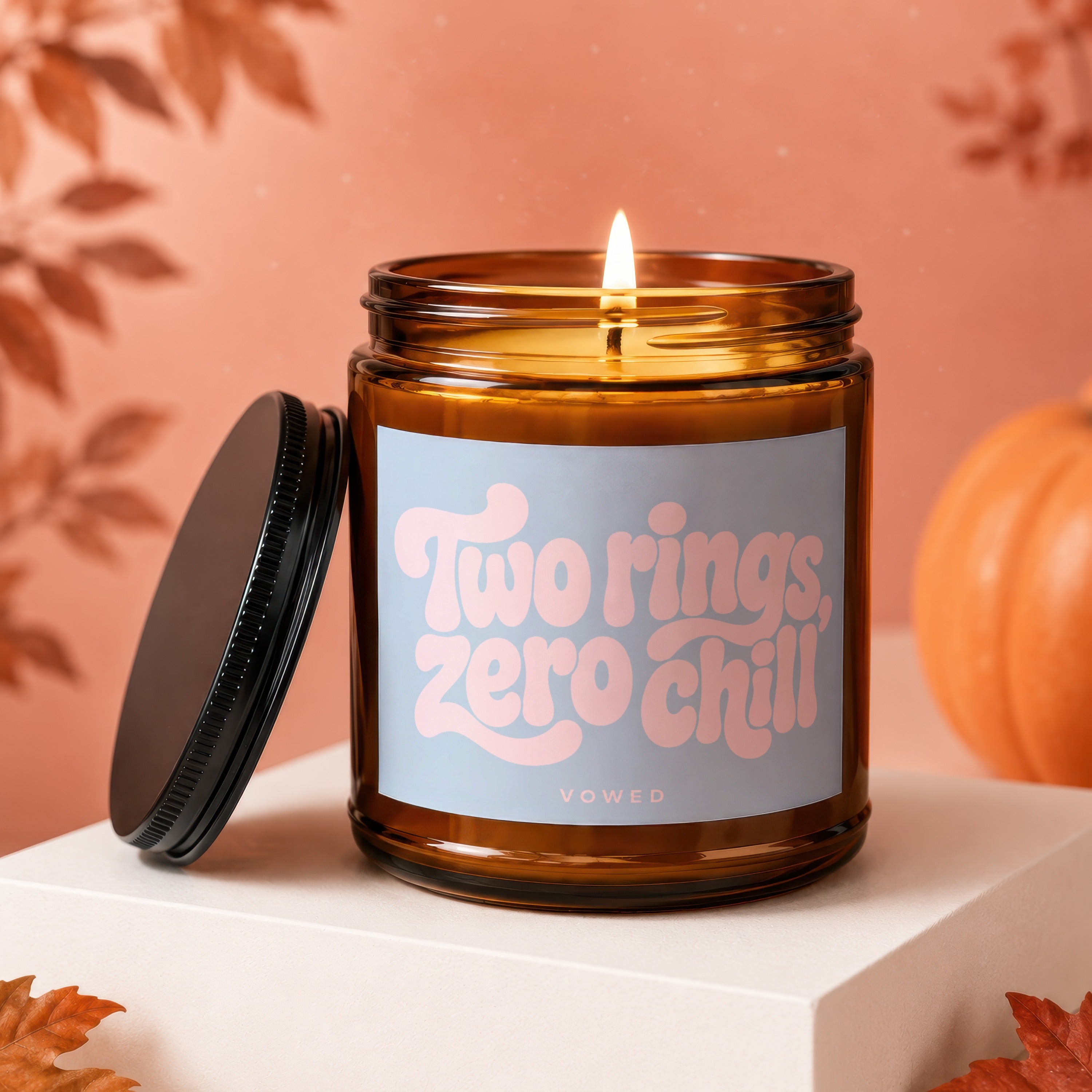 Amber jar candle with lit wick, "Two rings, zero chill" text, and pumpkin decoration.