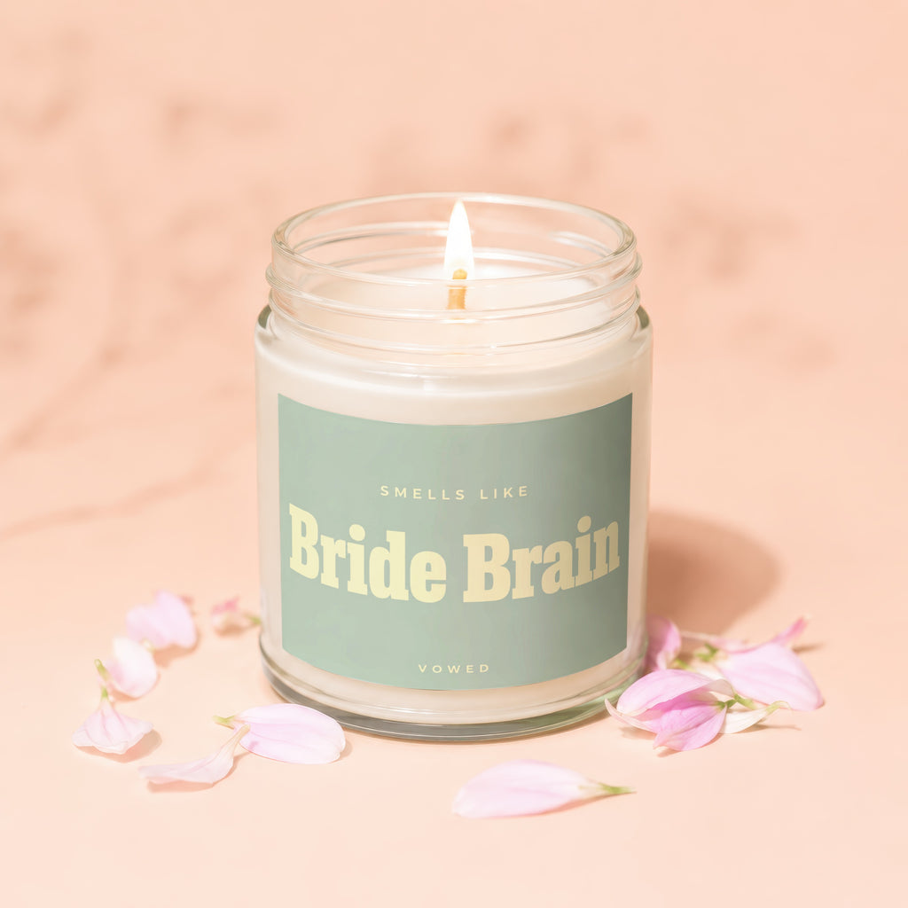 Glass jar candle with lit wick, "Smells Like Bride Brain" text, and pink petals.