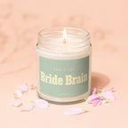 Glass jar candle with lit wick, "Smells Like Bride Brain" text, and pink petals.