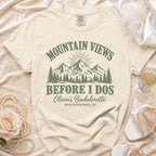 A cream-colored t-shirt with green text that reads Mountain Views Before I Dos, Olivia's Bachelorette, Breckenridge, CO, and a mountain graphic.