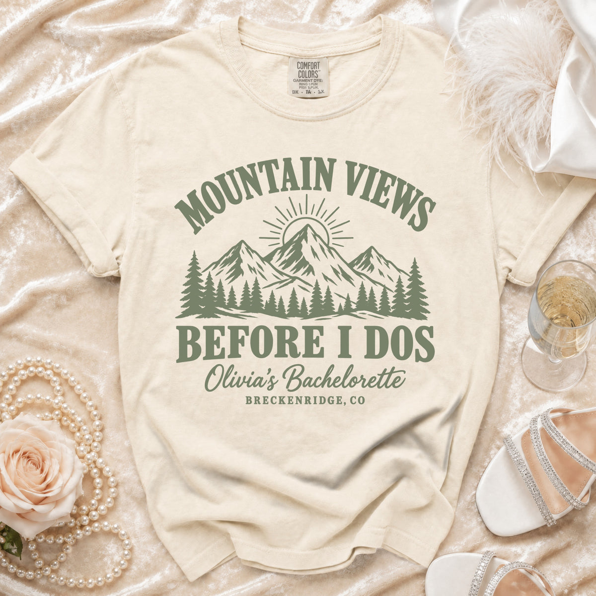 A cream-colored t-shirt with green text that reads Mountain Views Before I Dos, Olivia's Bachelorette, Breckenridge, CO, and a mountain graphic.