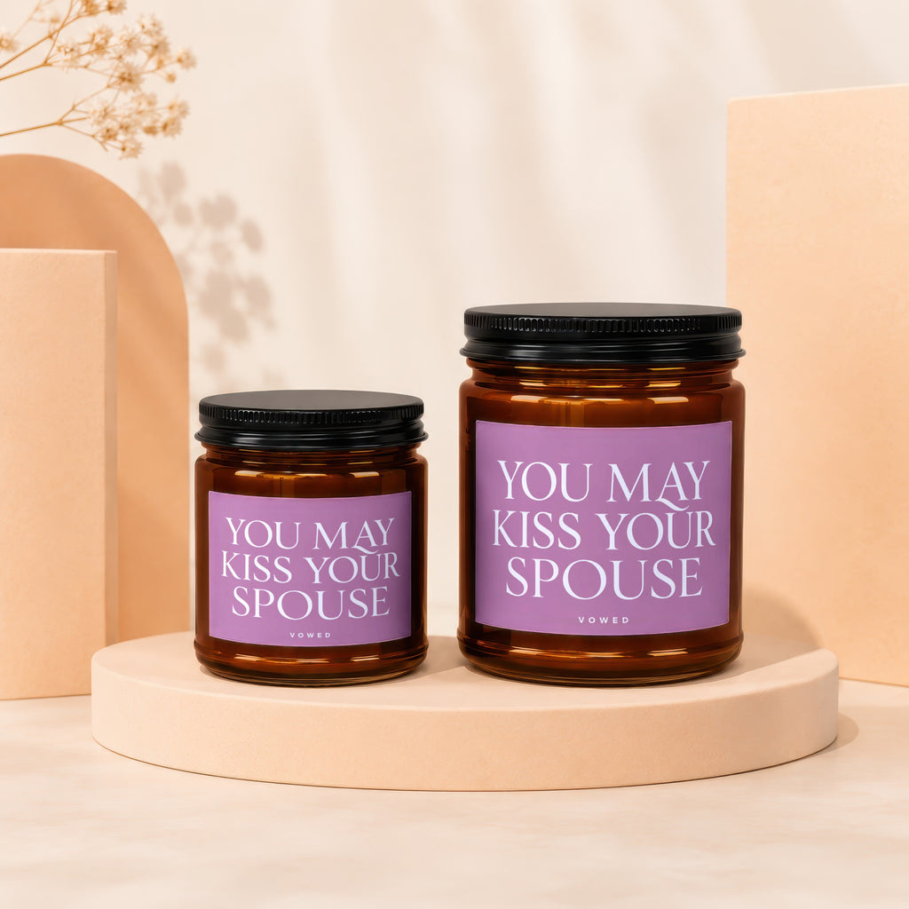 Two amber glass candles with black lids and purple labels reading "YOU MAY KISS YOUR SPOUSE".