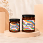 Two amber glass candles with colorful striped labels saying "Be Still, My Heart! You're Engaged!".