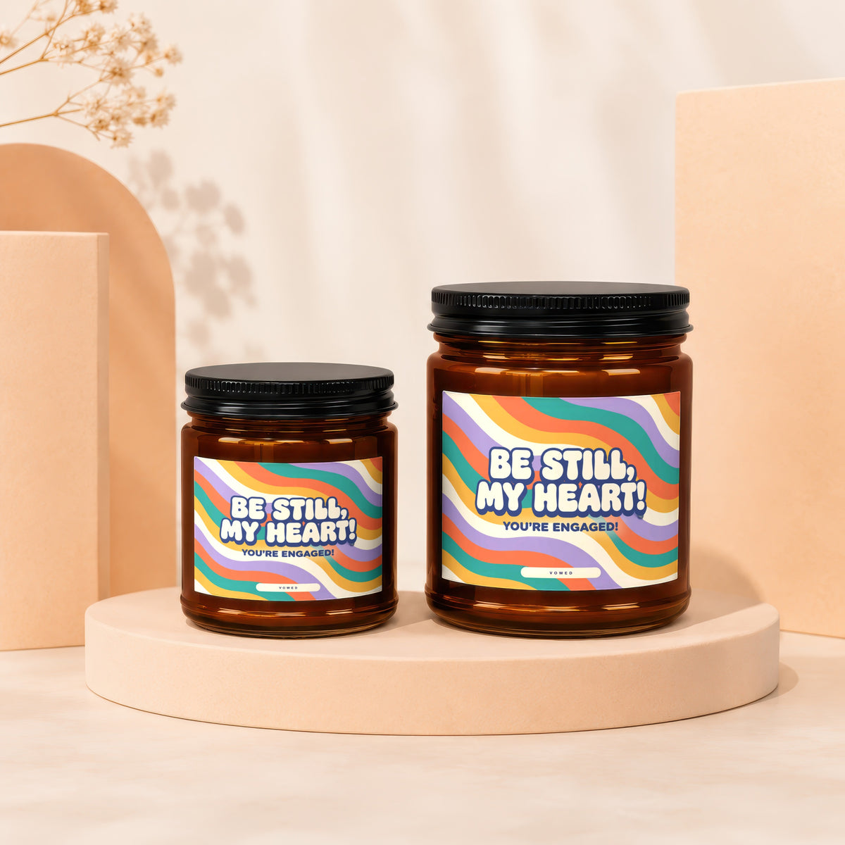 Two amber glass candles with colorful striped labels saying "Be Still, My Heart! You're Engaged!".
