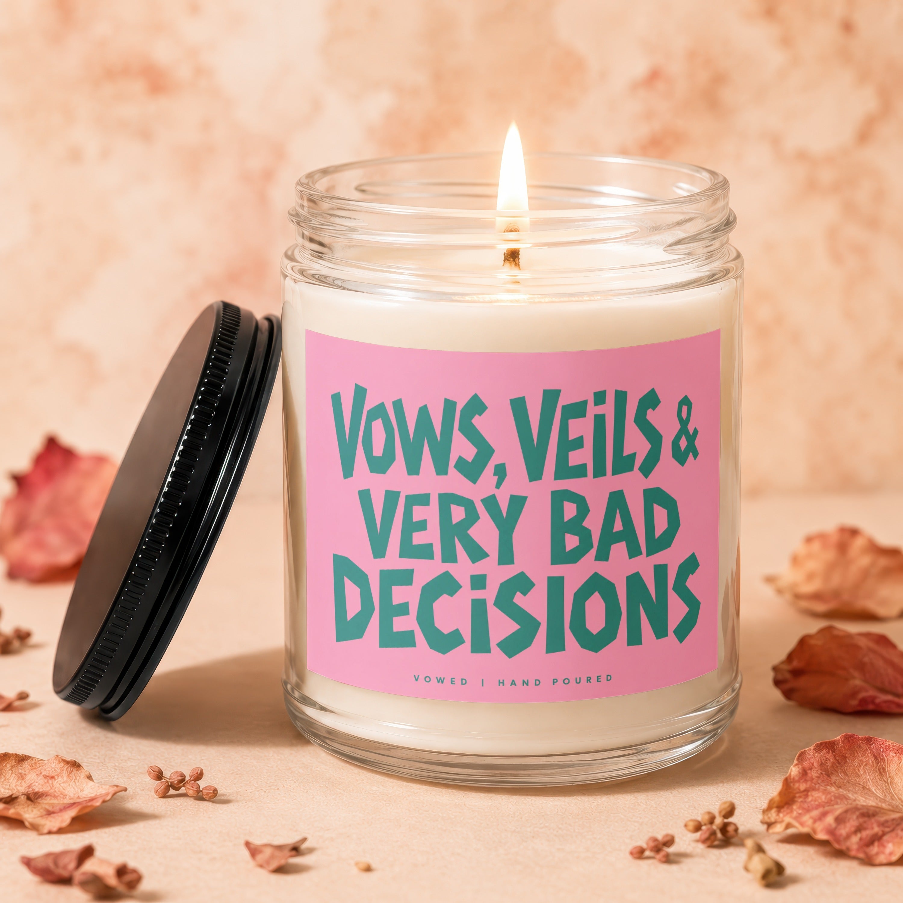 Glass jar candle with a lit wick and pink label saying "Vows, Veils & Very Bad Decisions".