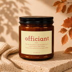 Amber candle with label reading "officiant noun, VOWED" on textured surface.