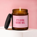 Lit candle in amber jar with pink label that reads "It's Giving Future Mrs."