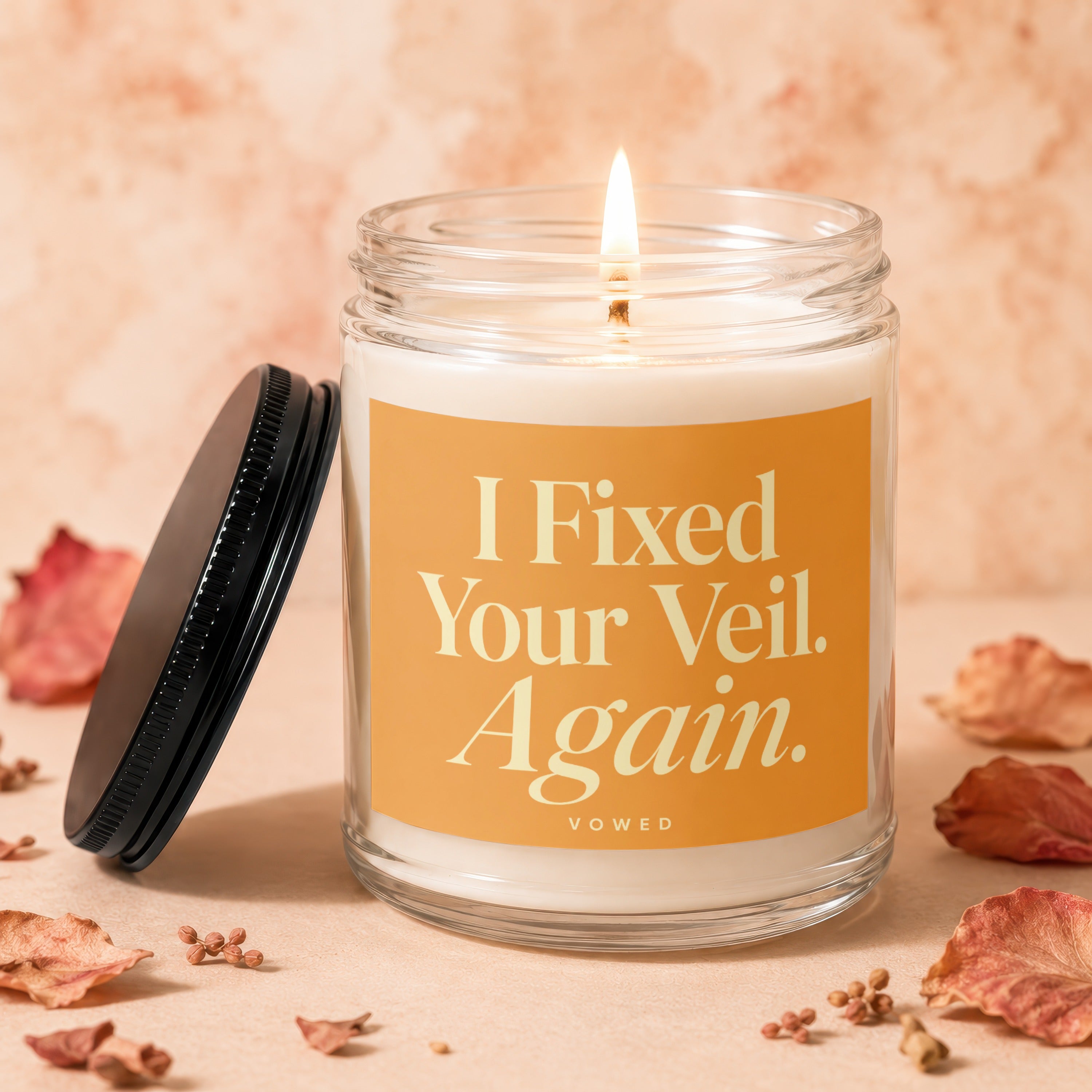 Glass candle with lit wick and orange label saying "I Fixed Your Veil. Again."