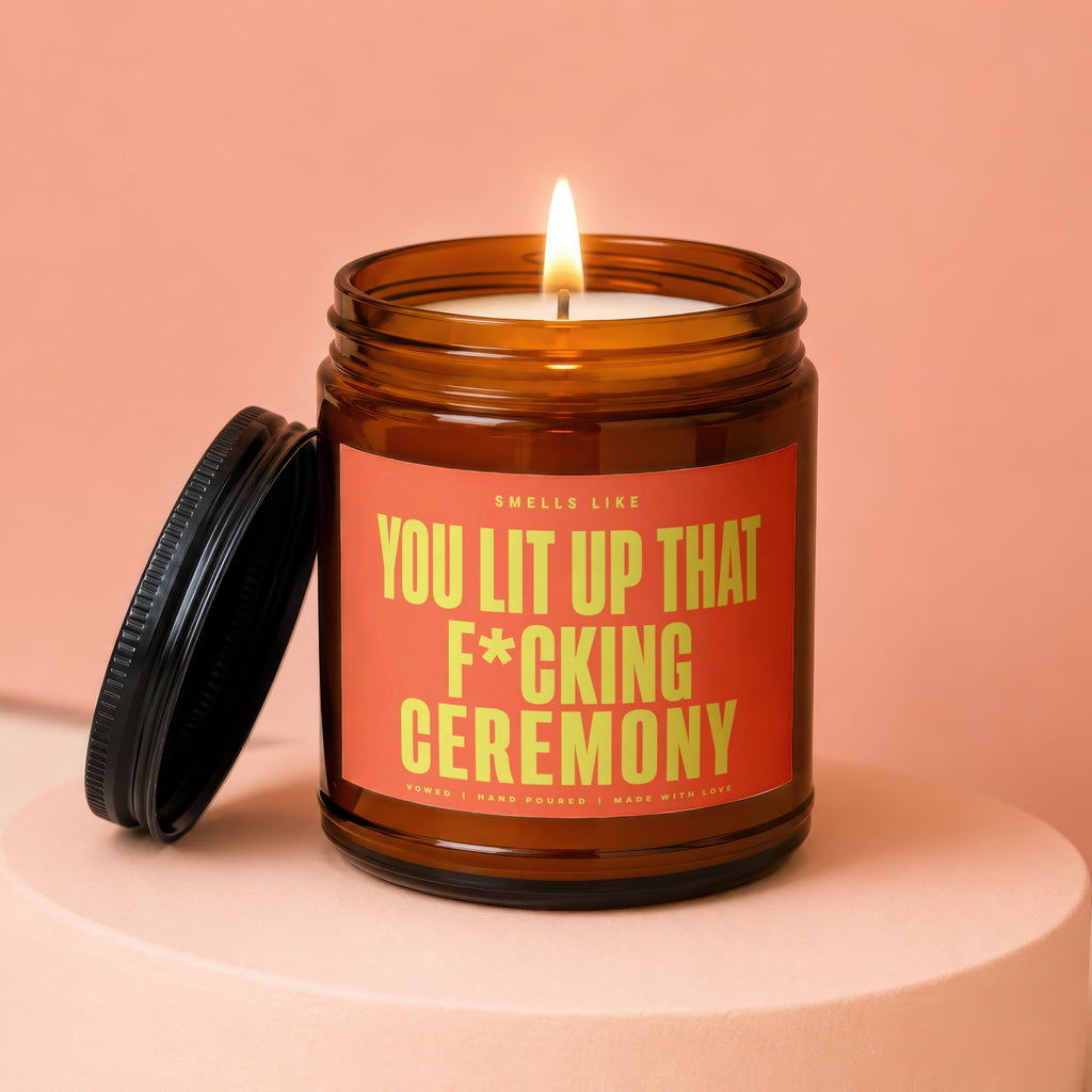 Amber jar candle with a lit wick and label stating "Smells Like You Lit Up That F*cking Ceremony."