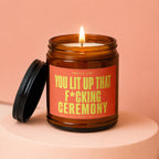 Amber jar candle with a lit wick and label stating "Smells Like You Lit Up That F*cking Ceremony."