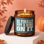 Amber candle with lit wick, lid removed, and text: Smells like HE FINALLY PUT A RING ON IT.