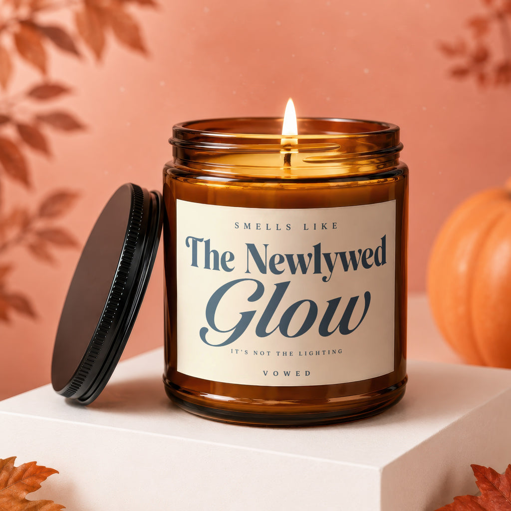 Lit amber glass candle with black lid, text "Smells Like The Newlywed Glow".