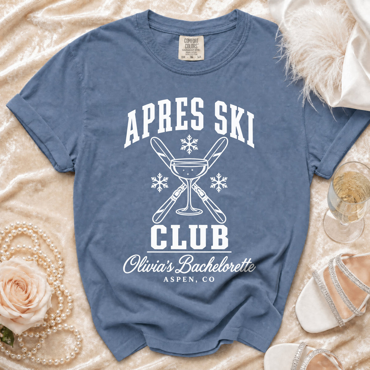Blue crewneck t-shirt featuring white retro text reading "APRES SKI CLUB OLIVIA'S BACHELORETTE ASPEN CO" with crossed skis, a martini glass, and snowflakes.