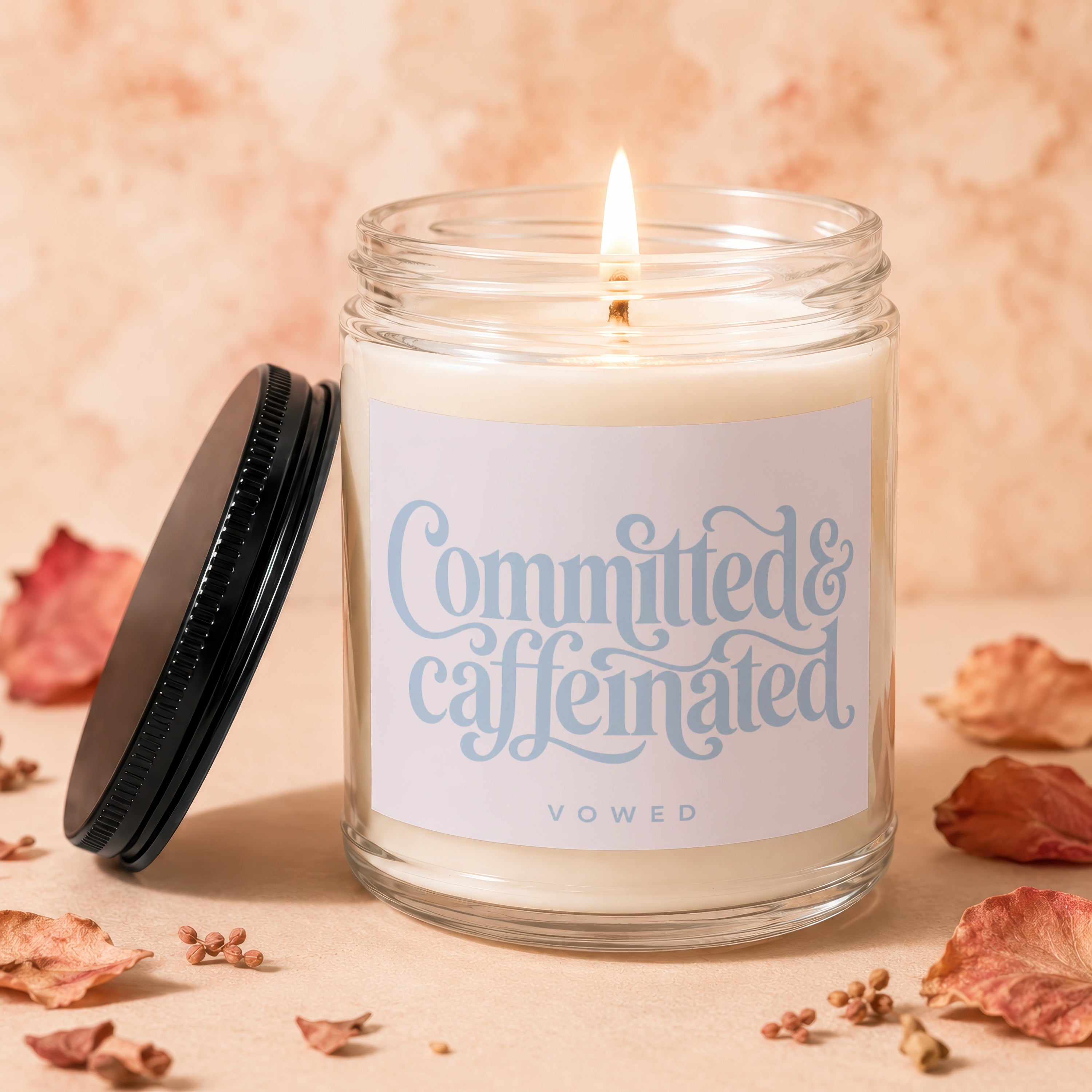 Lit candle with "Committed & Caffeinated Vowed" text on label, lid off and to the side.