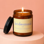 Amber jar candle with lit wick. Label reads "bridesmaid noun chief planner, problem solver, dress fluffer, and emotional support. will hold your bouquet, your secrets, and your drink. VOWED | HAND POURED".