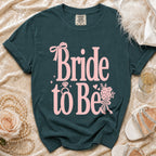 Dark teal t-shirt with pink "Bride to Be" text, ring, bow, and flowers.
