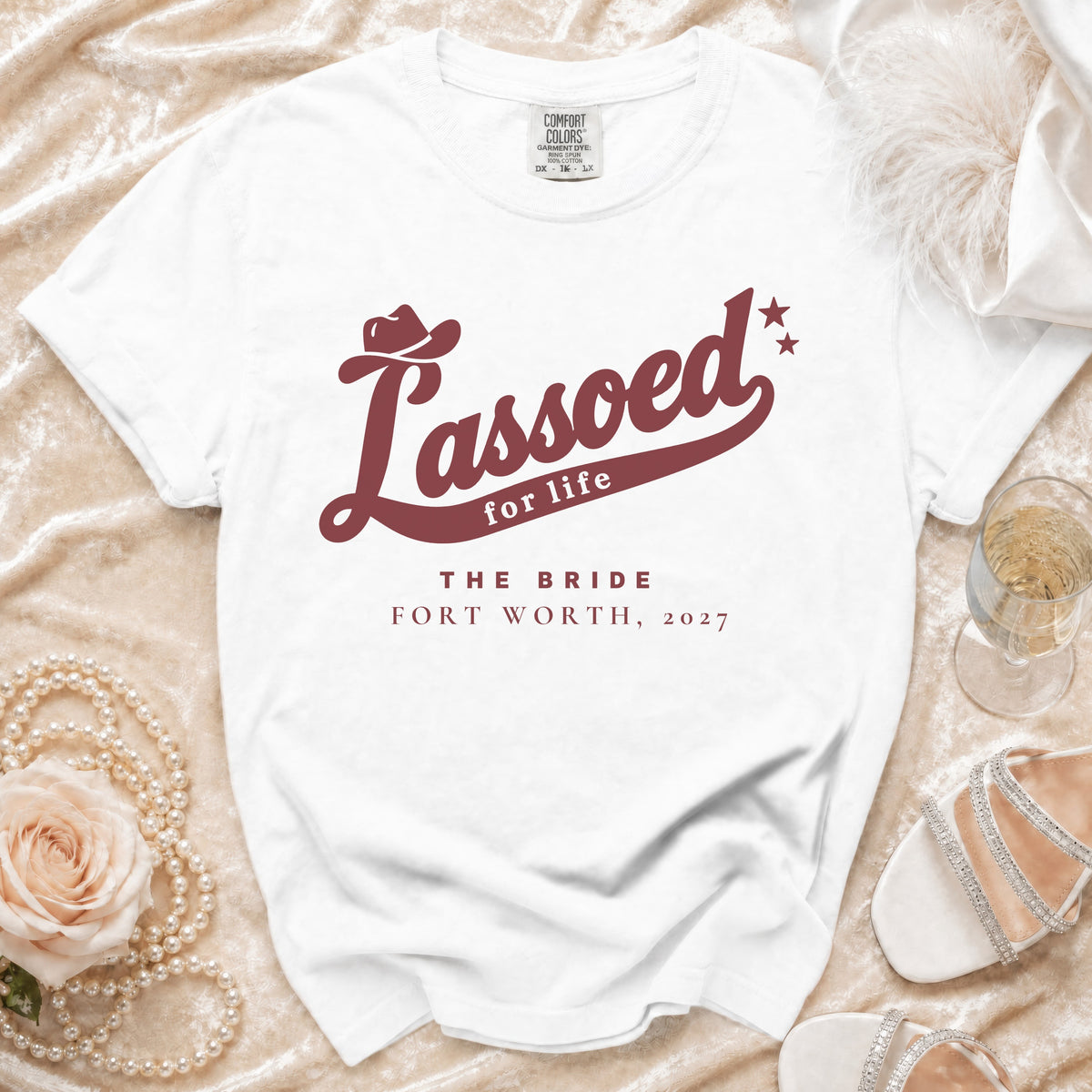 A white t-shirt featuring burgundy text that reads Lassoed for life, The Bride, Fort Worth, 2027.