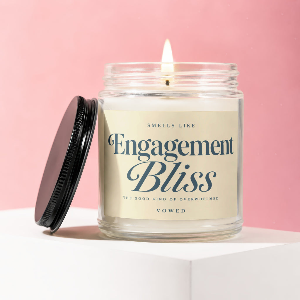 Lit candle in glass jar with "Engagement Bliss" label and black lid on a white surface.