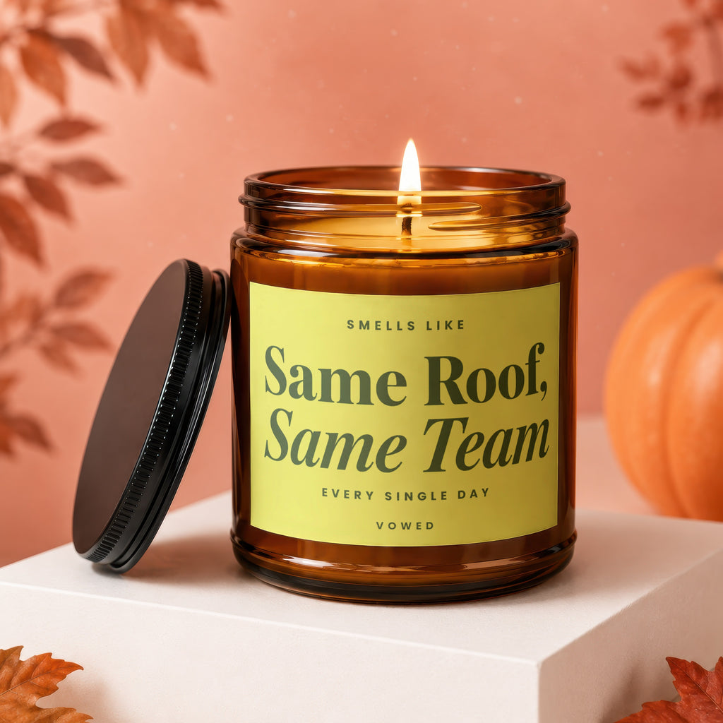 Lit amber candle with yellow label reading "Smells Like Same Roof, Same Team Every Single Day Vowed".