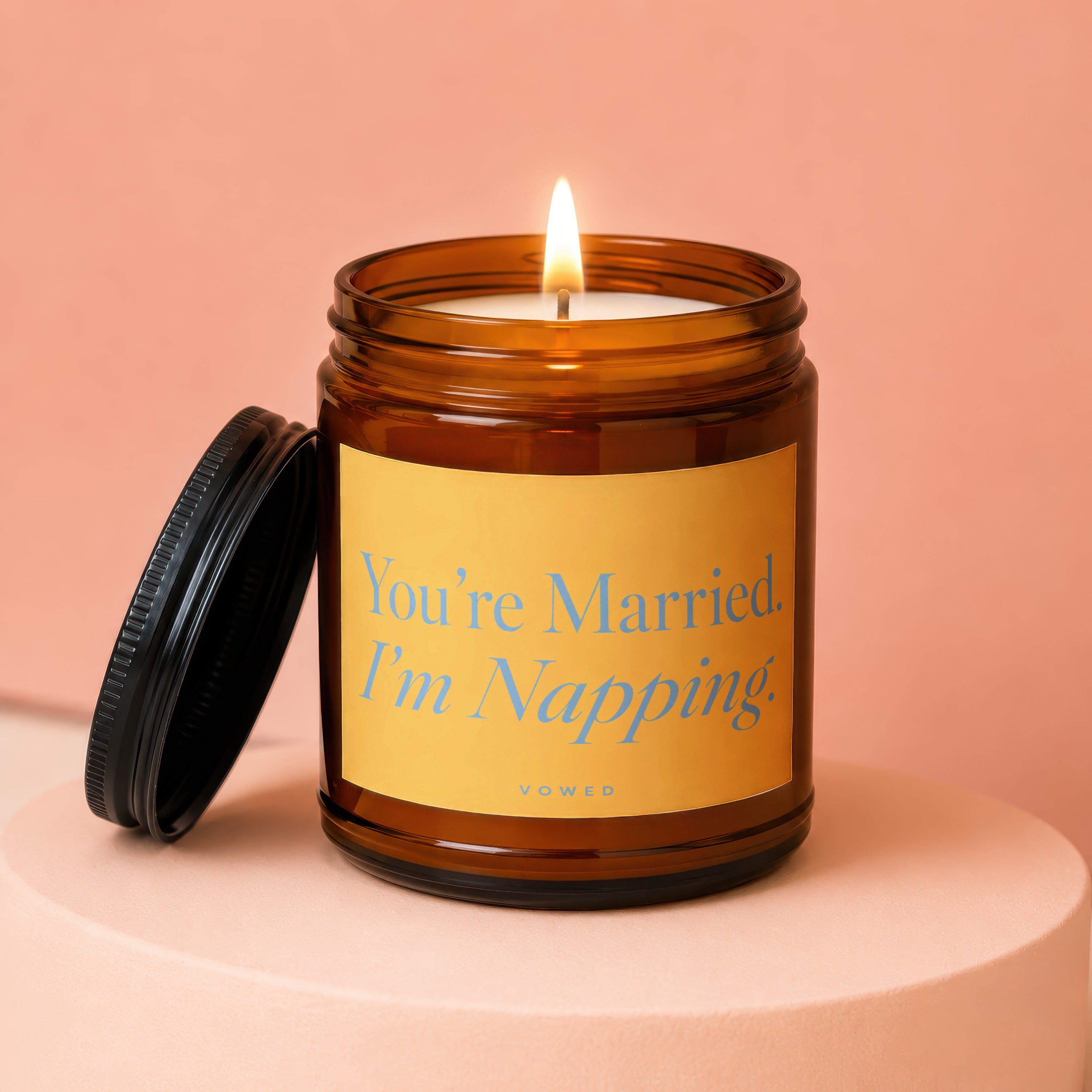 Lit amber glass candle with black lid and yellow label reading "You're Married. I'm Napping."