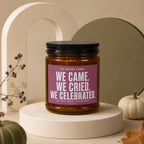 Amber jar candle with black lid, labeled "We Came. We Cried. We Celebrated."