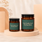 Two amber glass candles with green labels reading "Love Looks Good On You Both."