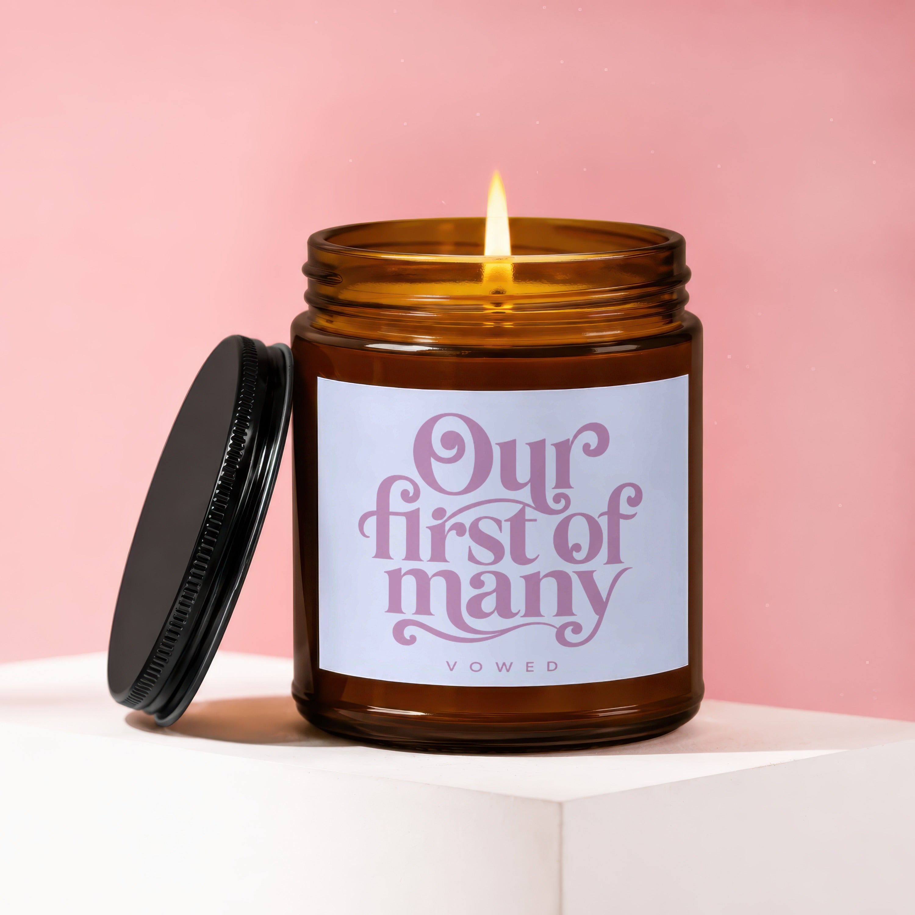 Amber glass candle with lit flame, lid off, reads "Our first of many VOWED."