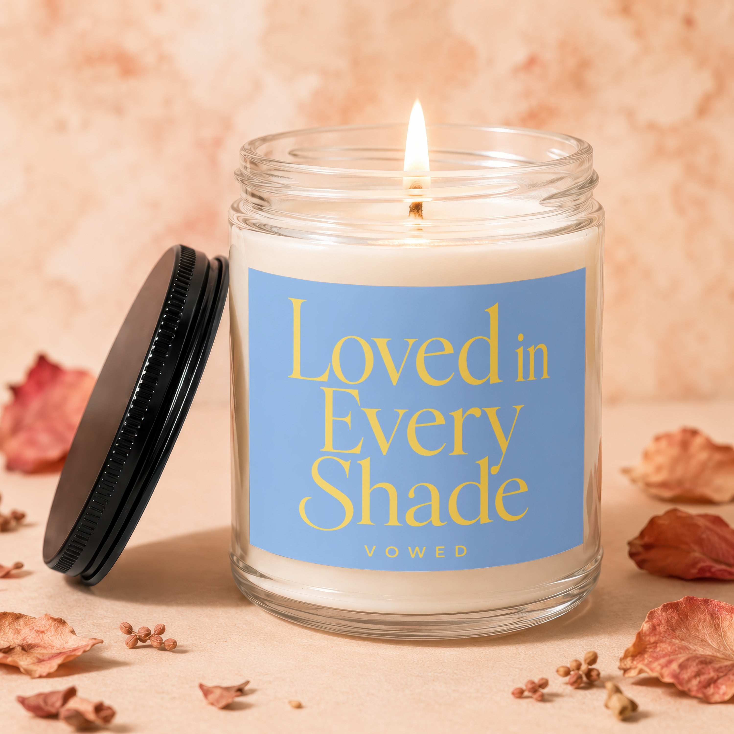 Lit candle in a glass jar with a blue label reading "Loved in Every Shade VOWED".