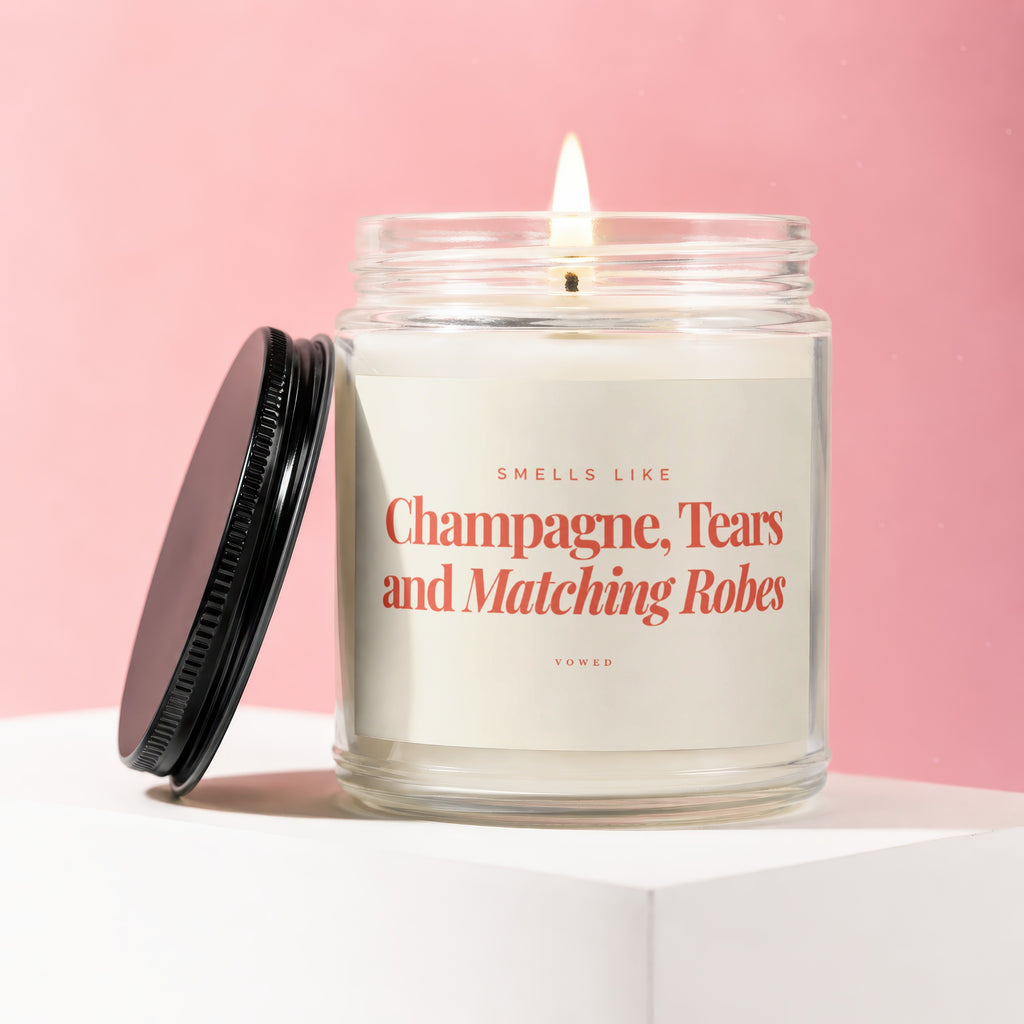 Candle with label "Smells Like Champagne, Tears and Matching Robes" on a pink background.