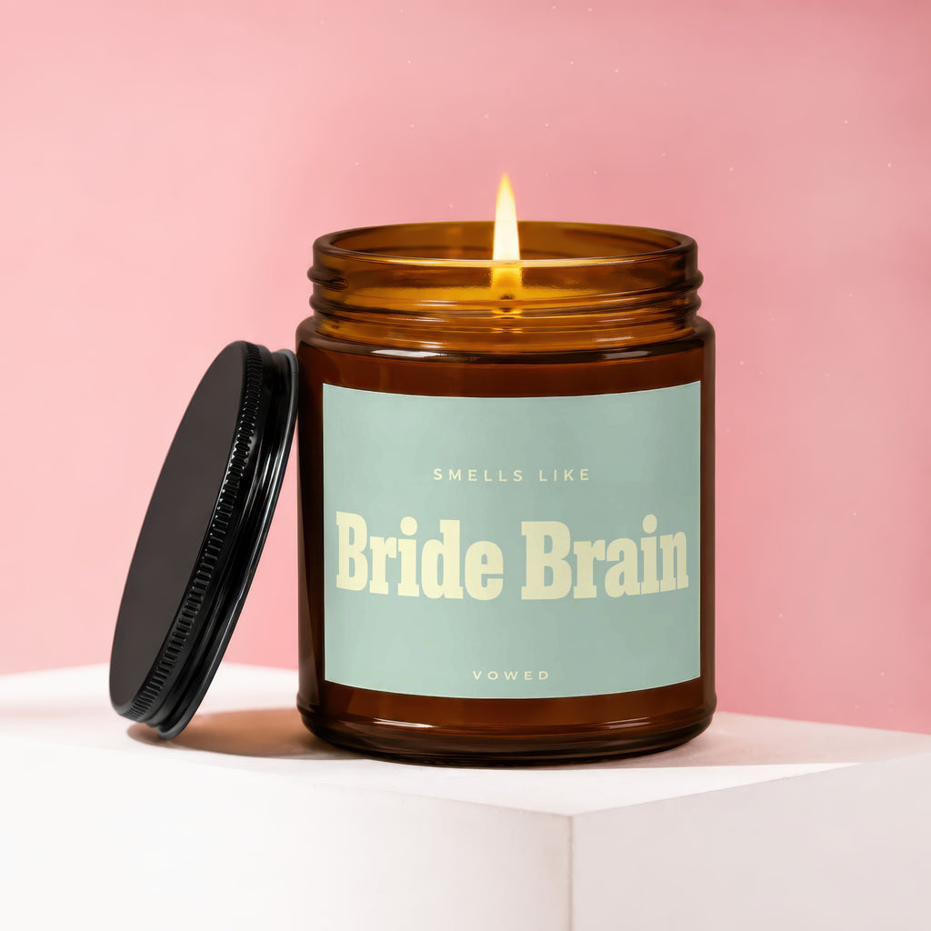 Amber glass candle with lid removed, lit wick, and label reading "Smells Like Bride Brain Vowed."