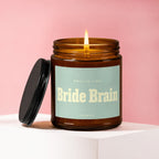 Amber glass candle with lid removed, lit wick, and label reading "Smells Like Bride Brain Vowed."