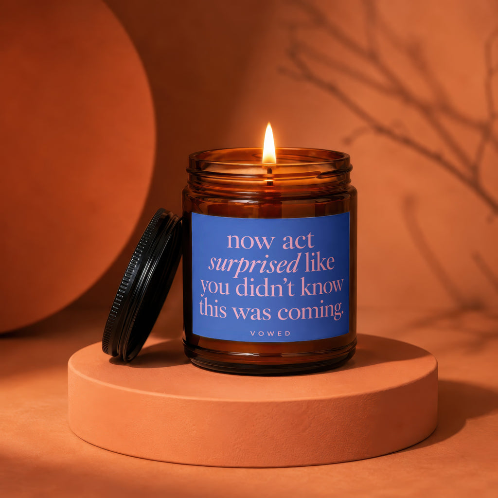 Amber glass candle with blue label reading "now act surprised like you didn't know this was coming."