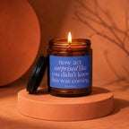Amber glass candle with blue label reading "now act surprised like you didn't know this was coming."