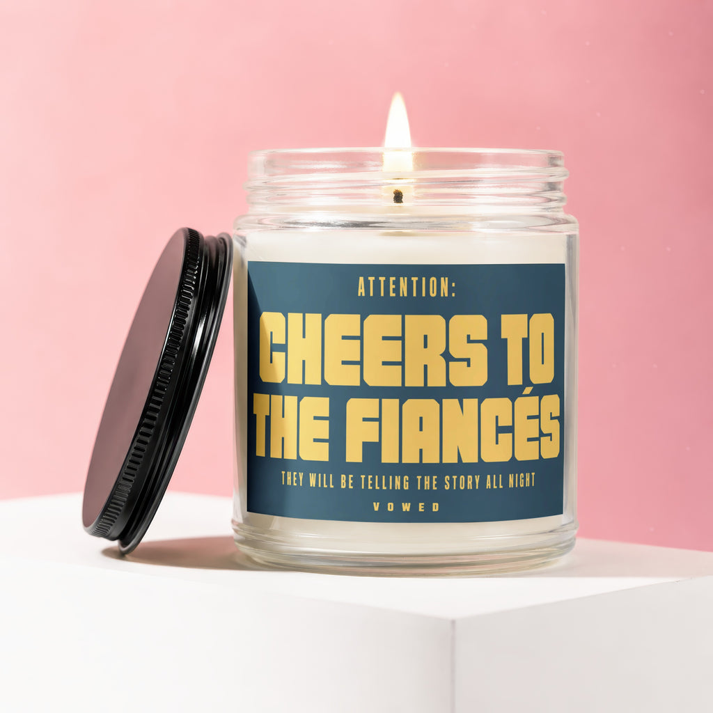 Glass candle with lid off, lit wick, reading "Cheers To The Fiances Vowed".