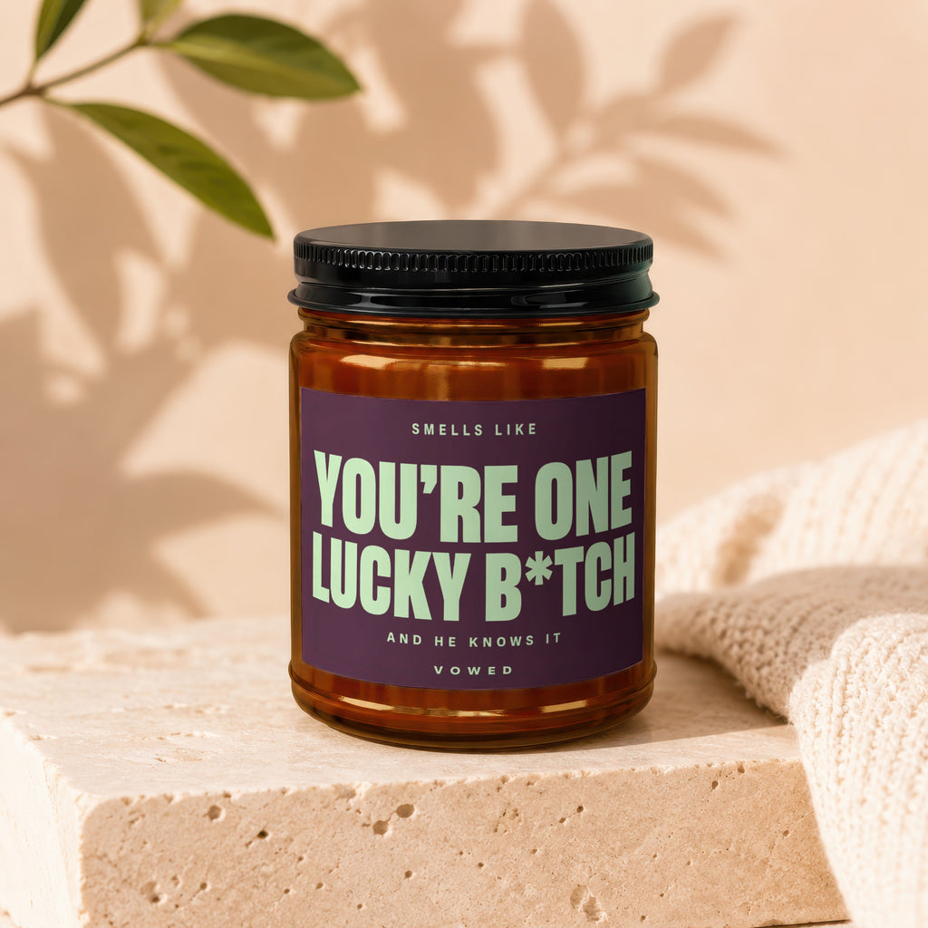 Amber jar candle with "You're One Lucky B*tch" text, on concrete block.