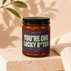 Amber jar candle with "You're One Lucky B*tch" text, on concrete block.