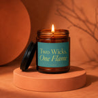 Lit amber glass candle with teal label reading "Two Wicks, One Flame".