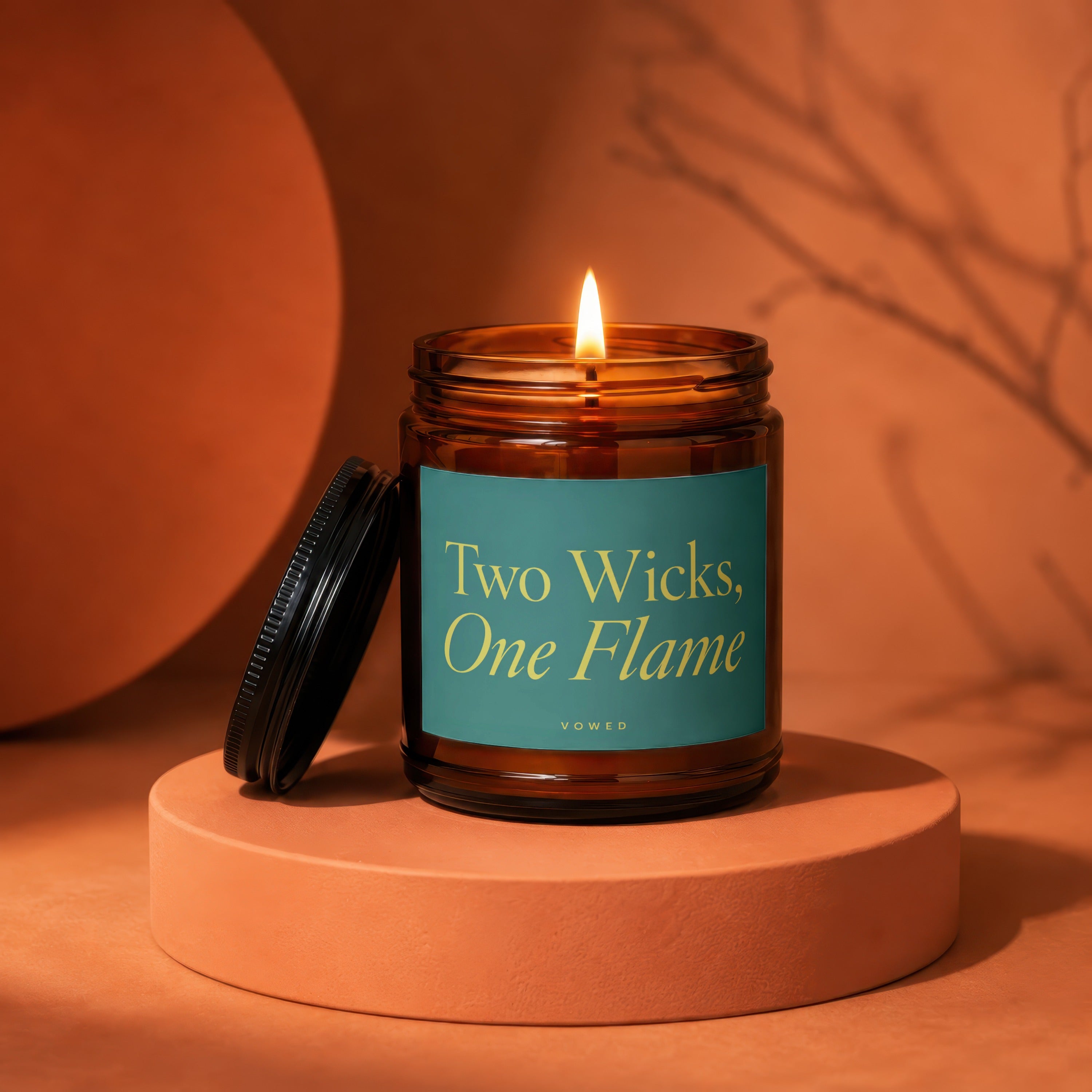 Lit amber glass candle with teal label reading "Two Wicks, One Flame".