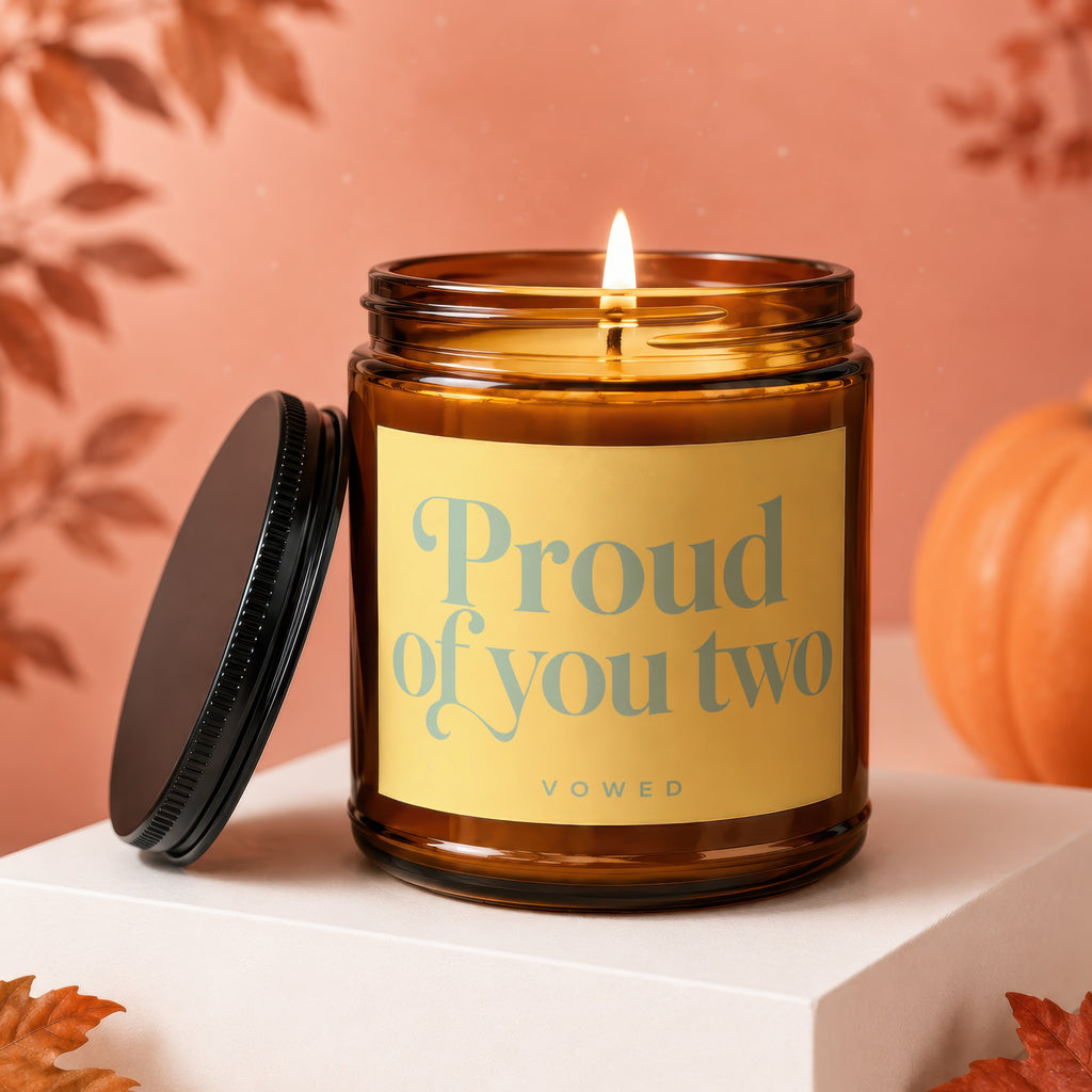Amber jar candle with lit wick, label reads "Proud of you two VOWED".