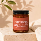 Amber glass candle with "Partners, officially VOWED" label on a cream textured surface.