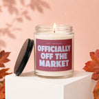 Lit candle in glass jar with "Officially Off The Market" text, next to its lid.
