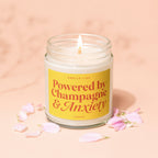 Candle with lit wick on yellow label reading "Smells Like Powered by Champagne & Anxiety."