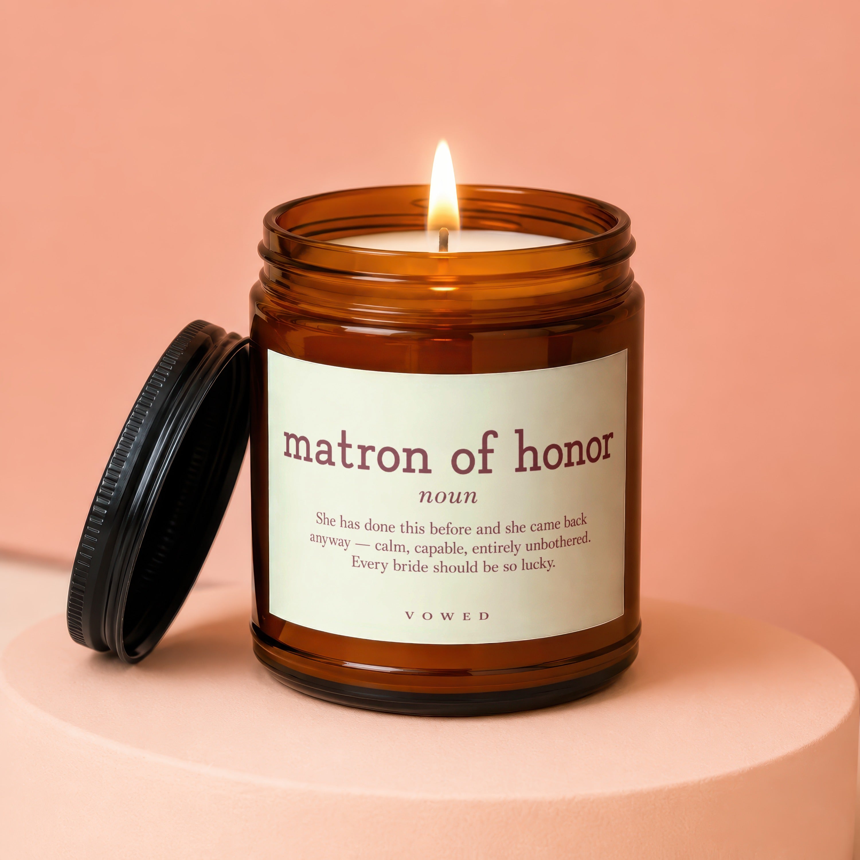 Amber jar candle labeled "matron of honor noun" with definition, burning at the top.