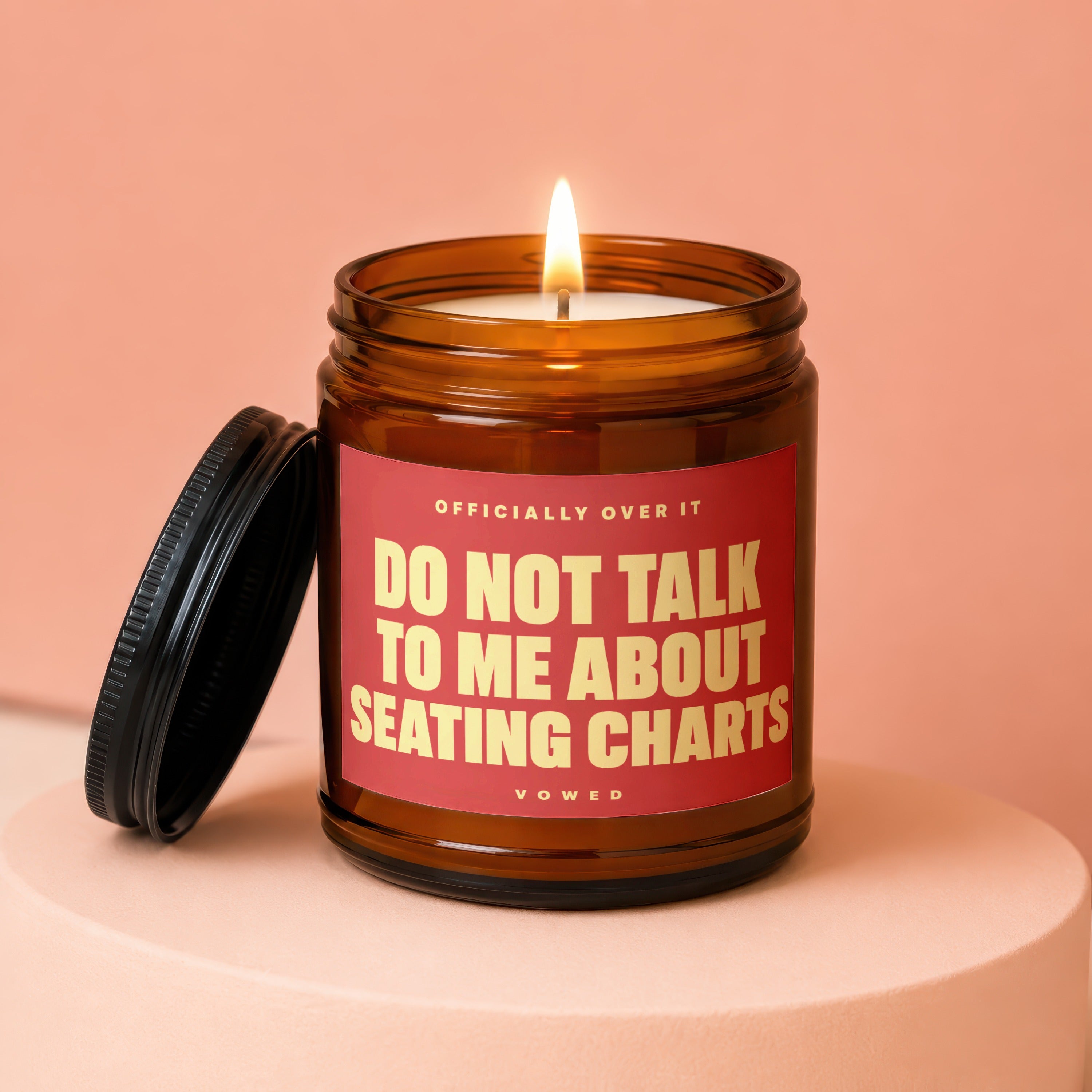 Amber jar candle with lit flame and red label reading "DO NOT TALK TO ME ABOUT SEATING CHARTS".
