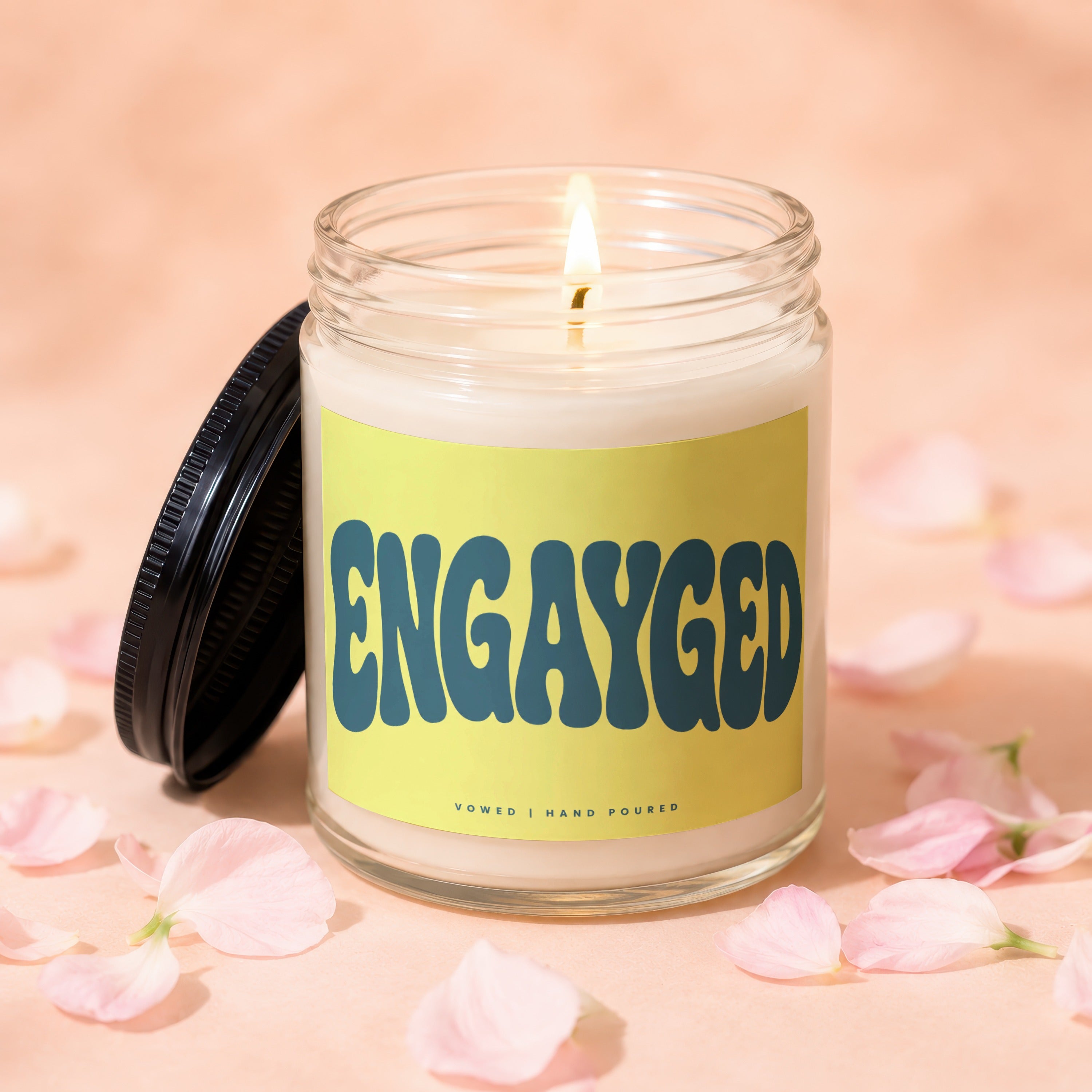 Glass candle with "ENGAYGED" text on yellow label, lit wick, and pink petals.