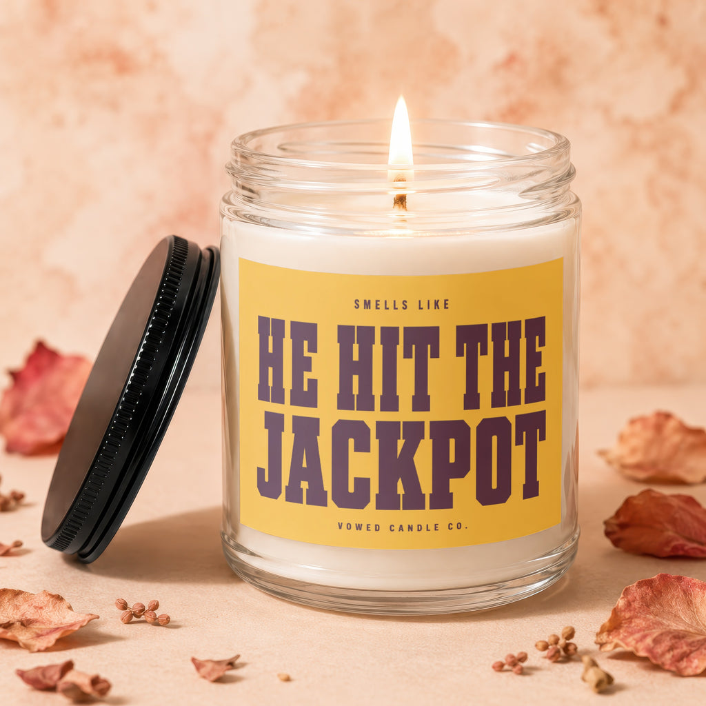Glass candle with lit wick, yellow label reading "HE HIT THE JACKPOT"