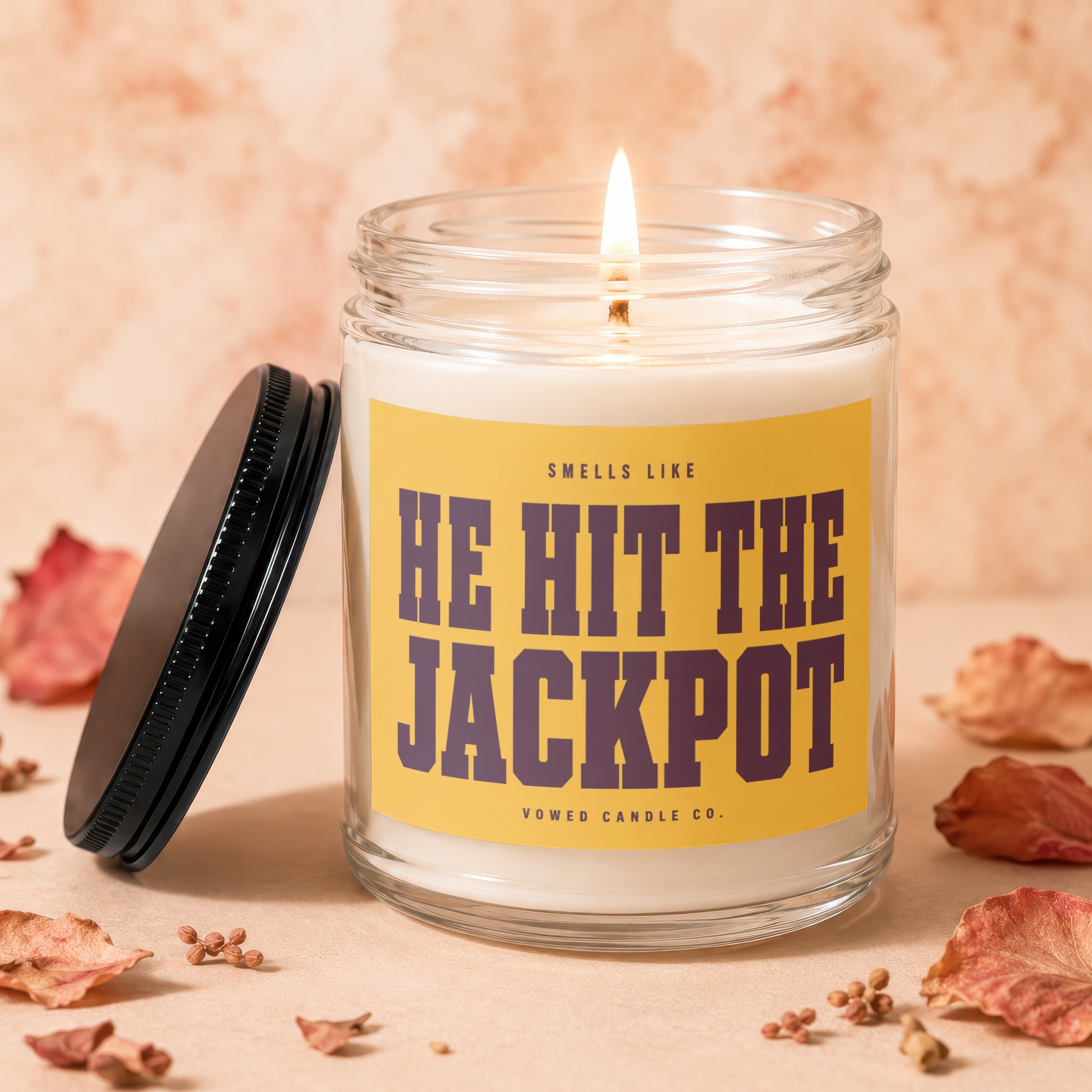 Glass candle with lit wick, yellow label reading "HE HIT THE JACKPOT"