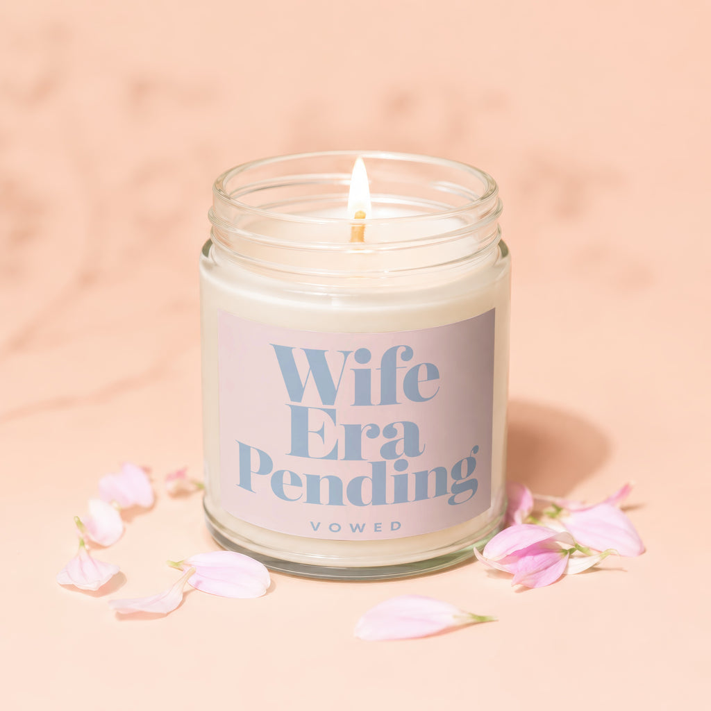 Lit candle in jar with "Wife Era Pending" text, surrounded by pink flower petals.