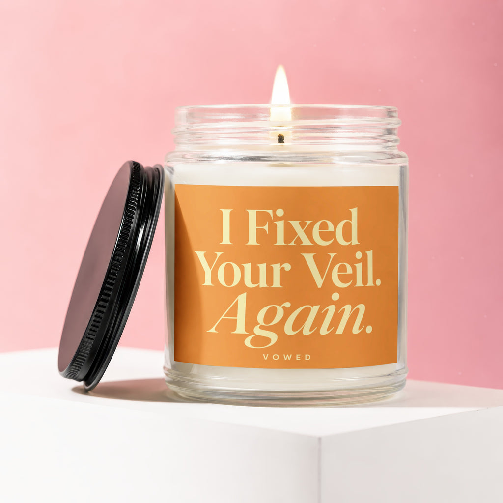 Glass jar candle with lit wick, orange label reads "I Fixed Your Veil. Again. VOwed."