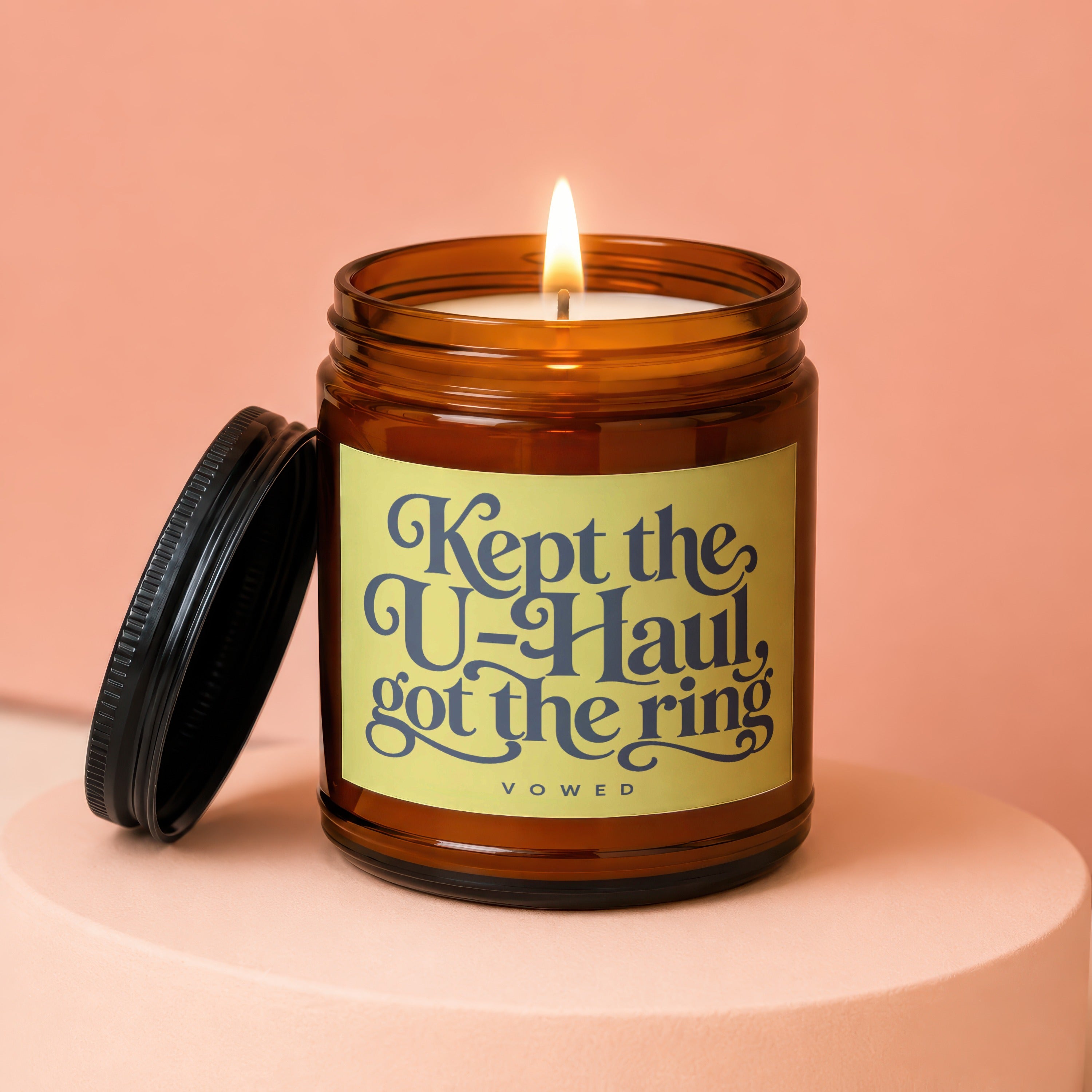 Amber jar candle with lit wick, reads "Kept the U-Haul, got the ring VOWED".